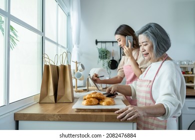 Asian Happy Family, Senior Mom And Daughter Cook Food And Sale At Home. Attractive Beautiful Girl And Elderly Older Mother Baking Croissant Then Packing Bakery Into Delivery Bag On Table In Kitchen.