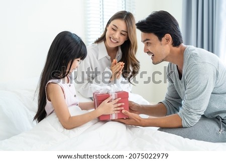 Asian Happy family, young parents surprise sleep daughter with gift. Attractive man and woman couple wake up their little kid from bed in bedroom to give Birthday present and celebrate tradition party