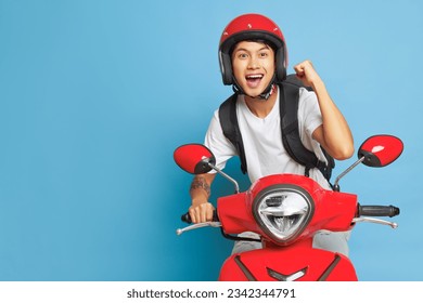 Asian happy guy in red helmet riding red motorbike and holds one fist up, posing on blue backdrop with wide smile, lifestyle concept, copy space - Powered by Shutterstock