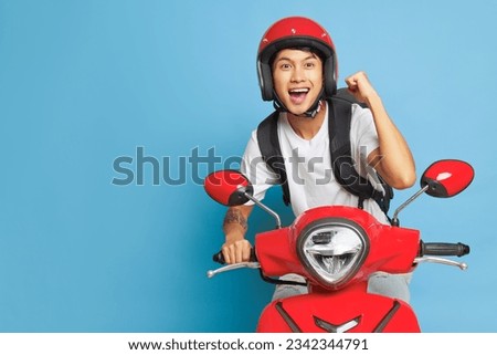 Asian happy guy in red helmet riding red motorbike and holds one fist up, posing on blue backdrop with wide smile, lifestyle concept, copy space