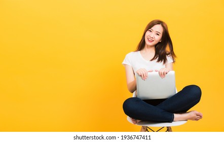 Asian Happy Portrait Beautiful Cute Young Woman Teen Smiling Sitting Crossed Legs On A Chair With Laptop Computer Looking To Camera Isolated, Studio Shot On Yellow Background With Copy Space