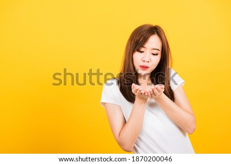 Asian happy portrait beautiful cute young woman teen standing wear t-shirt blowing kiss air something on hands looking to hand isolated, studio shot on yellow background with copy space