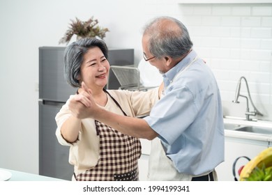 Asian Happy Senior Elderly Couple Standing In Kitchen At House Enjoy Retirement Life And Dancing Together. Loving Older Grandparents Look At Each Other And Smile. Relationship And Activity At Home.