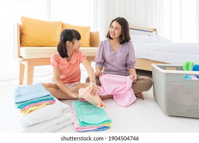 Asian Happy Smiling Mother And Daughter Sitting On The Floor In A Bedroom At Home While Helping Together Folding Clothes After Cleaning Clothing. Holiday And Family Concept.