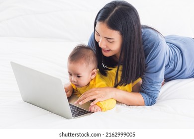 Asian Happy Young Mother And Baby Lifestyle Working On Laptop, Woman Mom And Daughter Reading And Learning Technology On Screen While Notebook Computer On Bedroom White Bed