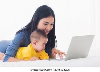 Asian Happy Young Mother And Baby Lifestyle Working On Laptop, Woman Mom And Daughter Reading And Learning Technology On Screen While Notebook Computer On Bedroom White Bed