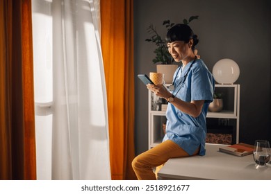 Asian healthcare professional in blue scrubs and stethoscope holds a coffee cup and engages with their smartphone, absorbing the morning sunlight streaming through the window by lively yellow curtains - Powered by Shutterstock