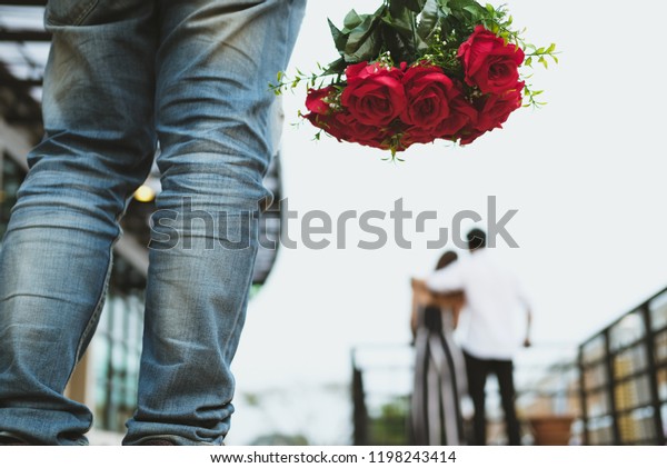 asian heartbroken man holding bouquet of red\
roses feeling sad while seeing woman dating with another man.\
broken heart, disappointed in love\
concept