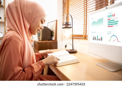Asian hijab woman read business books for work in the living room while business report graph showing on desktop computer in library. Portrait of Muslim girl reading book with staking books.