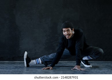 Asian Hip Hop Young Man With Tattoo Stretching Exercise His Legs On Floor Before Dance On Black Background. Attractive Man With Moustache Smiling Wear Knitted Hat, Ear Piercing Warm Up Body