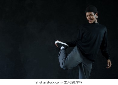 Asian Hip Hop Young Man With Tattoo Stretching Exercise His Leg Before Dance On Black Background. Attractive Man With Moustache Wear Knitted Hat, Ear Piercing, Tattoo On Face And Neck Warm Up Body