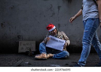 Asian Hopeless Poverty Man Wearing Christmas Hat Sitting On Wayside And Reading Newspaper About Economy With Hope. Aged Man With Beard Begging For Money, No Job. Society's Economic Problem.