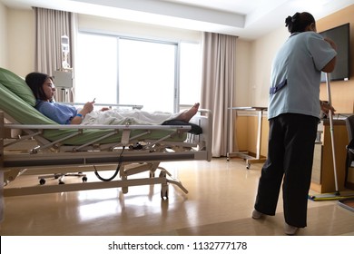 Asian Hospital Room Service Cleaner Woman Are Cleaning Floor With Mop In  Patient Room While Young Patient Woman Lying In Hospital Bed And Using Smart Mobile Phone While Recovering In Hospital