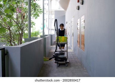 Asian Hotel Maid In Blue Uniform Push Housekeeping Cart On The Corridor In Front Of The Hotel Room. The Laundry Basket, Glass Cleaner, Rubber Gloves, Door Mat And Vacuum Cleaner Are On The Cart.