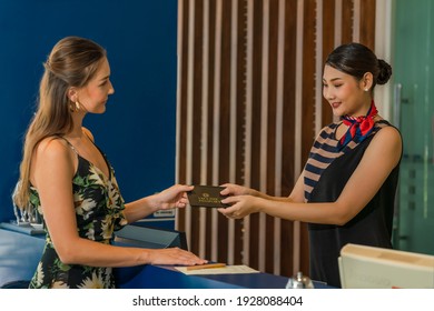 Asian Hotel Reception Or Airline Checkin Officer Receive Vaccine Passport From Caucasian Woman Guest Or Passenger As Regulation For Traveller During Coronavirus Covid-19 Pandamic