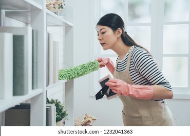 Asian Housekeeper In Apron Dusting The Bookshelf By Feather Duster Taking Up The Picture Frame Carefully Cleaning In Living Room At Home. Young Wife In Rubber Gloves Doing Housework.