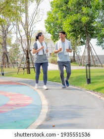 Asian Housewife And Her Husband Are Jogging In The Green Park. Happy Middle Aged Couple Exercising In The Evening. People Are Enjoying Life Doing Sports Together. Family Leisure Togetherness Concept.