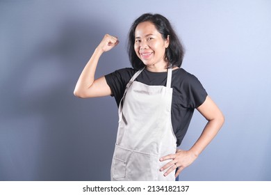 Asian housewife woman chef looks happy wearing apron and raising clenched fists, tensing muscles, feeling strong and full of energy isolated over grey background. Copy space. - Shutterstock ID 2134182137