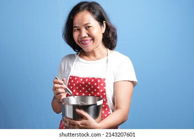 Asian housewife woman chef looks happy wearing apron holding cooking utensil and pointing up, standing isolated over blue background. Copy space.