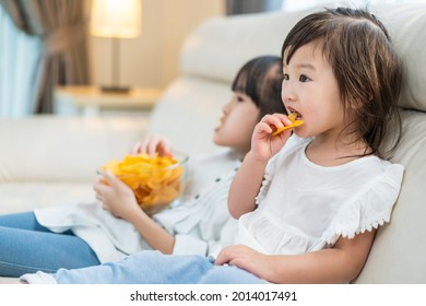 Asian Hungry Little Girl Sibling Sisters Puts Snack In Mouth With Hand. Two Sweet Preschool Kid Resting On Sofa At Home Enjoy Eat Fast Food, Potato Chips. Tasty And Unhealthy Food For Children Concept