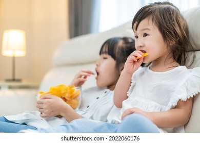 Asian Hungry Little Girl Sibling Sisters Puts Snack In Mouth With Hand. Two Sweet Preschool Kid Resting On Sofa At Home Enjoy Eat Fast Food, Potato Chips. Tasty And Unhealthy Food For Children Concept