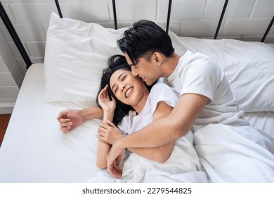 Asian husband kissing his wife while lying down sleep on bed in bedroom. Beautiful new marriage couple in pajamas feeling happy and relax spending leisure time in the early morning together in house. - Powered by Shutterstock