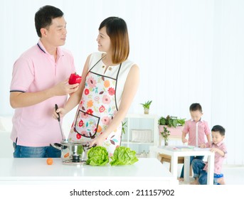 Asian Husband And Wife Cooking In The Kitchen And Kids Are Doing Schoolwork In The Living Room