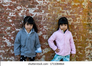 Asian Identical Female Twins Child Wearing Jacket During Cold Weather And Standing On An Old Brick Wall Background