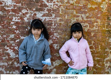 Asian Identical Female Twins Child Wearing Jacket During Cold Weather And Standing On An Old Brick Wall Background