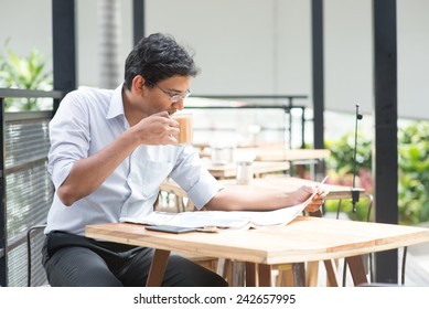 Asian Indian Business Man Reading Newspaper While Drinking A Cup Hot Milk Tea During Lunch Hour At Cafeteria.