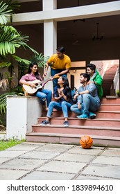 Asian Indian college students playing music with guitar while sitting in campus on stairs or over lawn