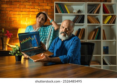 Asian Indian elderly couple with disappointed expressions reviewing their retirement investments on a laptop, concerned about the drop in stock market returns during a late-night session at home - Powered by Shutterstock