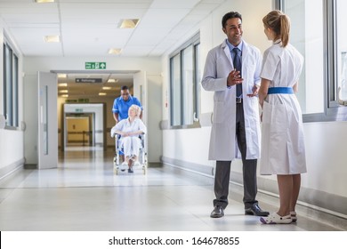 Asian Indian Male Doctor & Female Nurse In Hospital Corridor With A & Senior Female Wheelchair Patient And African American Nurse In Background