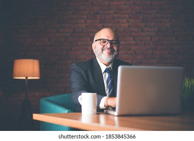 Asian Indian Senior Financial Businessman Sitting At His Workstation Or Desk In Front Of A Computer, Laptop And Tablet. Speaking On Phone While Doing Some Paperwork