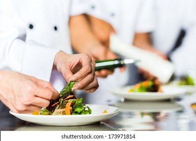 Asian Indonesian Chef Along With Other Cooks In Restaurant Or Hotel Commercial Kitchen Cooking, Finishing Dish Or Plate