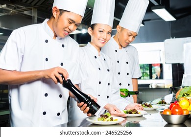 Asian Indonesian Chef Along With Other Cooks In Restaurant Or Hotel Commercial Kitchen Cooking, Finishing Dish Or Plate