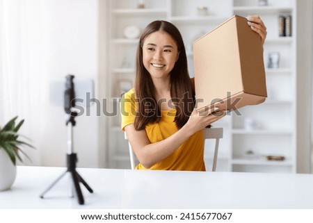 Asian influencer woman making video review of product at camera, smiling korean female holding cardboard box, recording content for her blog, sitting at desk at home and using smartphone on tripod