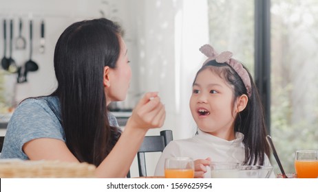 Asian Japanese Family Has Breakfast At Home. Asian Mom And Daughter Happy Talking Together While Eating Bread, Drink Orange Juice, Corn Flakes Cereal And Milk On Table In Modern Kitchen In Morning.