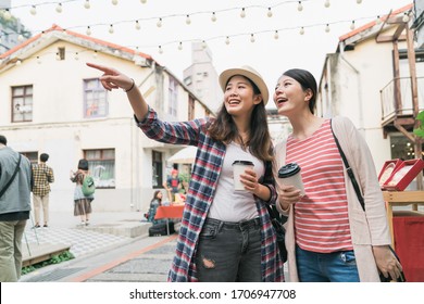 Asian Japanese Female Tourists Walking Between Rows On Creative Market. Young Travel Woman Point Finger Showing Friend With Special Thing While Shopping In Outdoor Holiday Bazaar. Ladies Drink Coffee