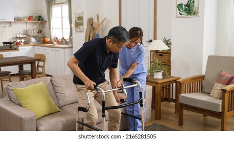 Asian Japanese Older Male Stroke Patient Practicing Using A Walker With The Assistance Of His Personal Care Attendant In The Living Room At Home