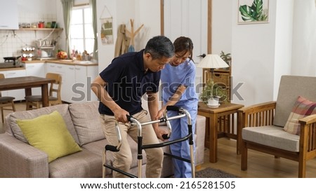 asian Japanese older male stroke patient practicing using a walker with the assistance of his personal care attendant in the living room at home