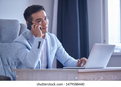 Asian Kazakh Businessman In A Suit And Glasses With A Laptop Computer In The Office Speaks By Mobile Phone, Solves Business Issues, Negotiates A Deal.