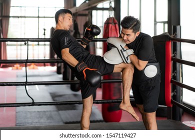 Asian Kick Boxer Strike With The Right Knee To Professional Trainer In Boxing Studuim In The Background At Fitness Gym. Boxer Are Hitting The Sandbag For Bodybuilding And Healthy Lifestyle Concept.
