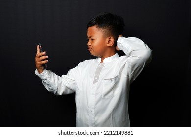 Asian Kid Become Confused And Scratching The Head While Watch On The His Cell Phone Screen Isolated On Black Background. Side View Close Up Details.