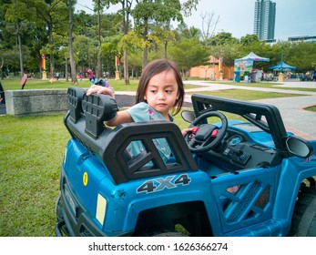 An Asian Kid Driving Electric Toy Car In A Tropical Park. Outdoor Toys. Children In Battery Power Vehicle. Little Girl Riding Toy Truck In The Garden. Family Playing In The Backyard.