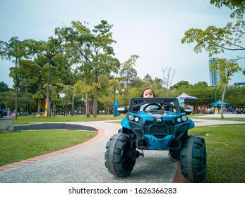 An Asian Kid Driving Electric Toy Car In A Tropical Park. Outdoor Toys. Children In Battery Power Vehicle. Little Girl Riding Toy Truck In The Garden. Family Playing In The Backyard.