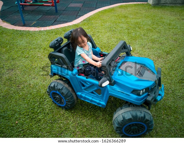 An\
Asian kid driving electric toy car in a tropical park. Outdoor\
toys. Children in battery power vehicle. Little girl riding toy\
truck in the garden. Family playing in the\
backyard.