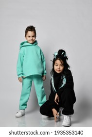 Asian Kid Girl In Black Stylish Hoodie And Pants Sits Squatted At Caucasian Kid Boy Standing In Pastel Modern Green, Mint Color Sports Suits Over White Background. Friendship And Funky Time