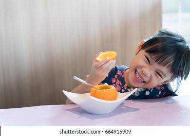 Asian Kid Girl Happy Eating Fresh Orange. Healthy Lifestyle. Kid Sitting In A Restaurant At Table Eating Oranges Background Empty Copy Space.