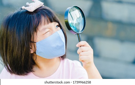 Asian Kid Girl Looking Through A Magnifying Glass On Nature Park Wearing Fabric Mask.Eye Care,Healthcare, Education, Science.Genius Kid Girl Learning In School Finding.Reopen School From Covid-19.